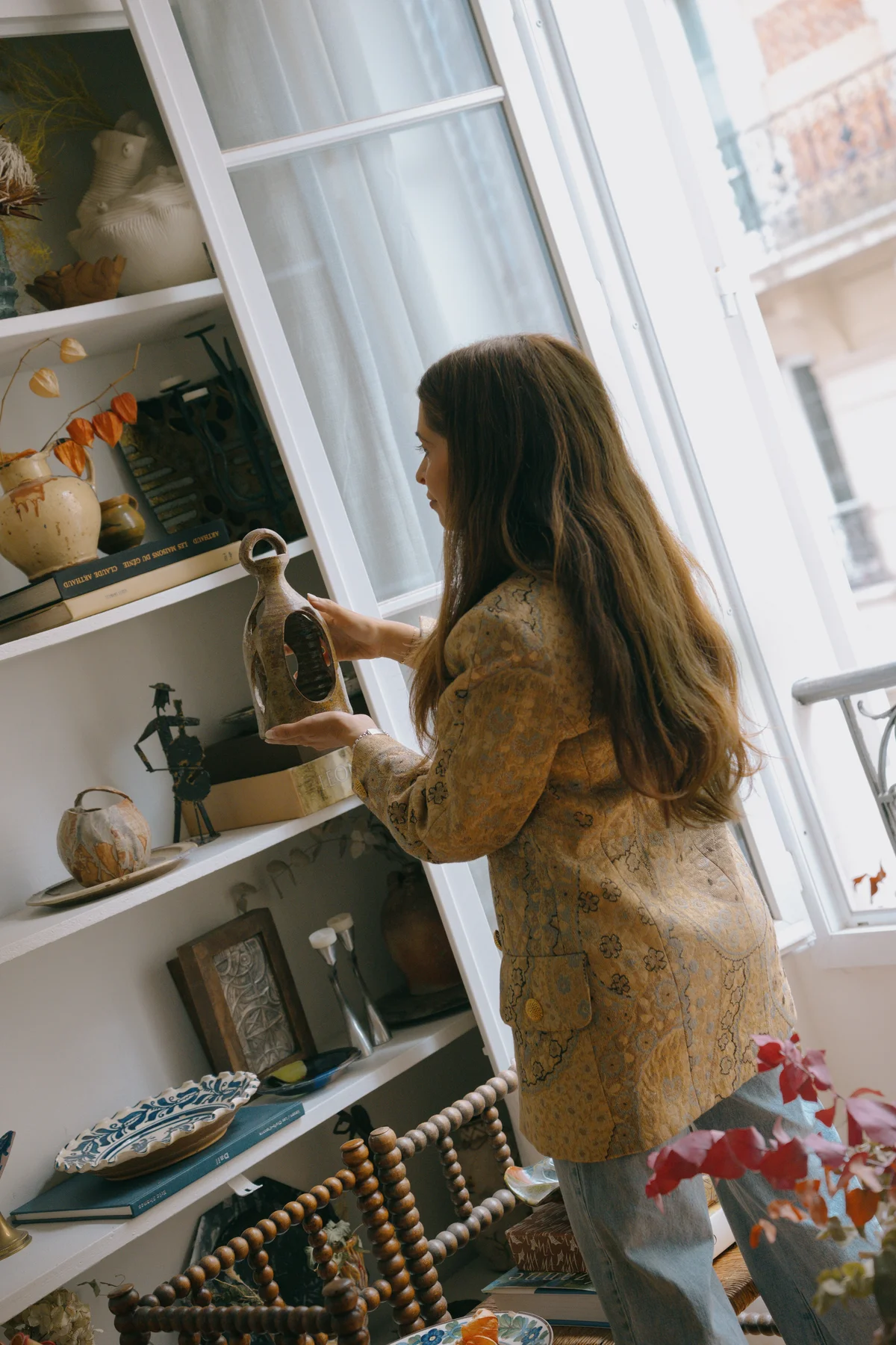 Javaneh Pirooz reaching up to place a glazed vessel on a white shelving unit of ceramics and books, at a tall window with a wrought-iron balcony and a pale stone facade beyond.