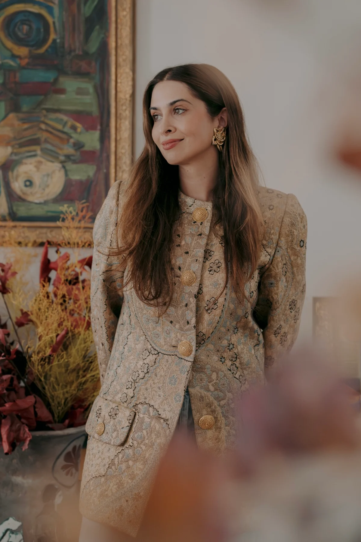 Javaneh Pirooz in an embroidered brocade coat, looking away from the camera, before a large modernist painting with dried foliage in a painted urn beside her.