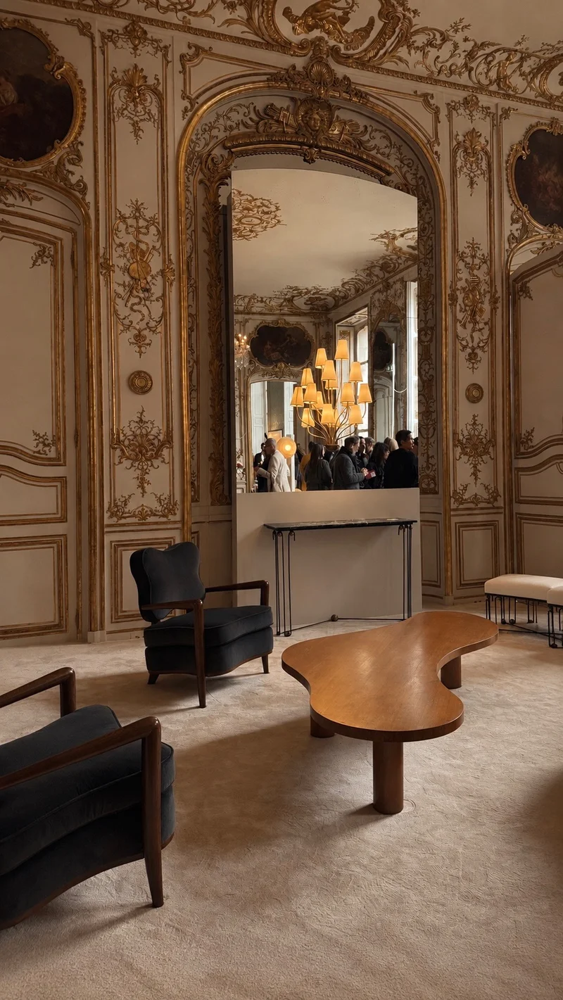 A gilded salon: carved and gilt boiserie panelling with painted overdoors, a tall arched mirror reflecting a chandelier and figures beyond, a kidney-shaped timber low table and dark velvet armchairs on a pale carpet.