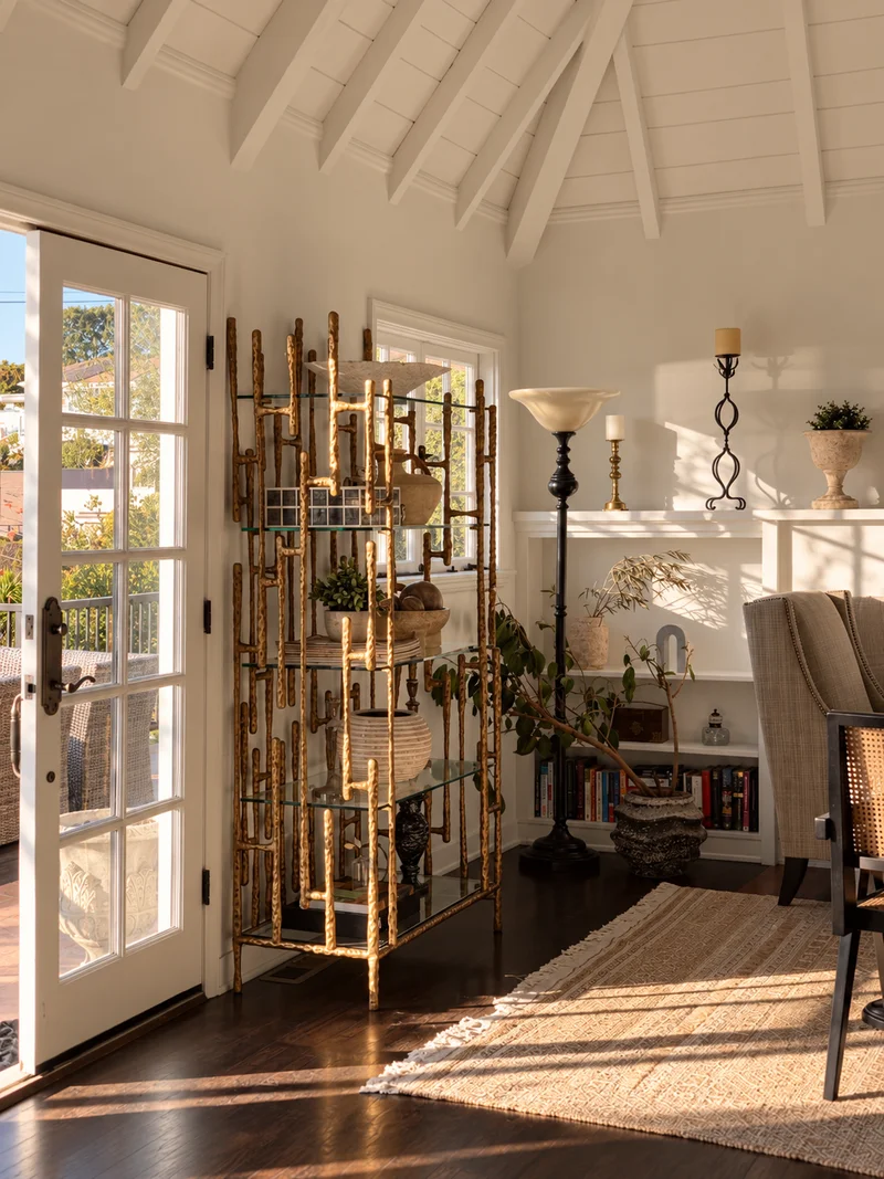 A brass etagere of books and objects beside a glazed door in a tall white-beamed room, floor lamps and a wing chair beyond, on a jute rug.