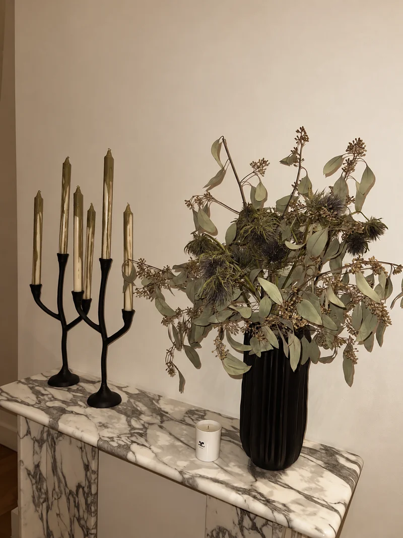 A black candelabra and a fluted black vase of dried eucalyptus and seed heads on a veined marble console against a bare plaster wall.