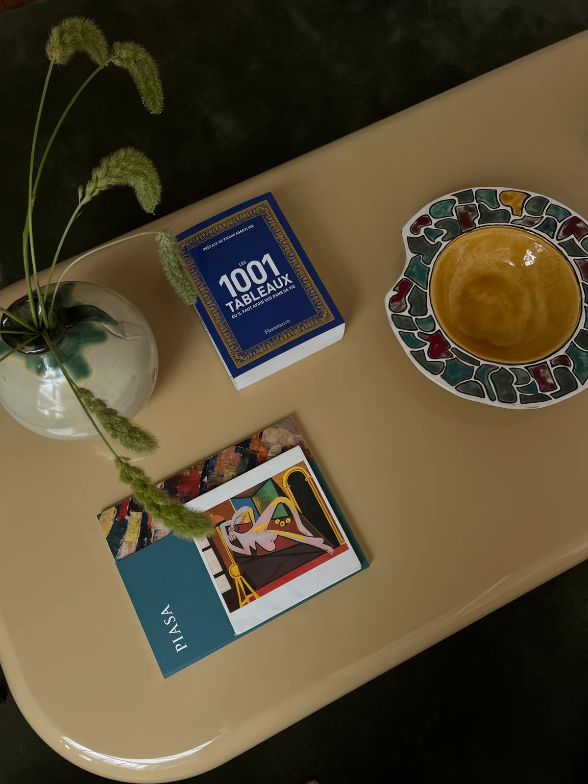 Looking down onto the cream table: a glass bulb vase holding one grass stem, a stack of art books, and a hand-painted ceramic dish with an amber-glazed bowl set into it.
