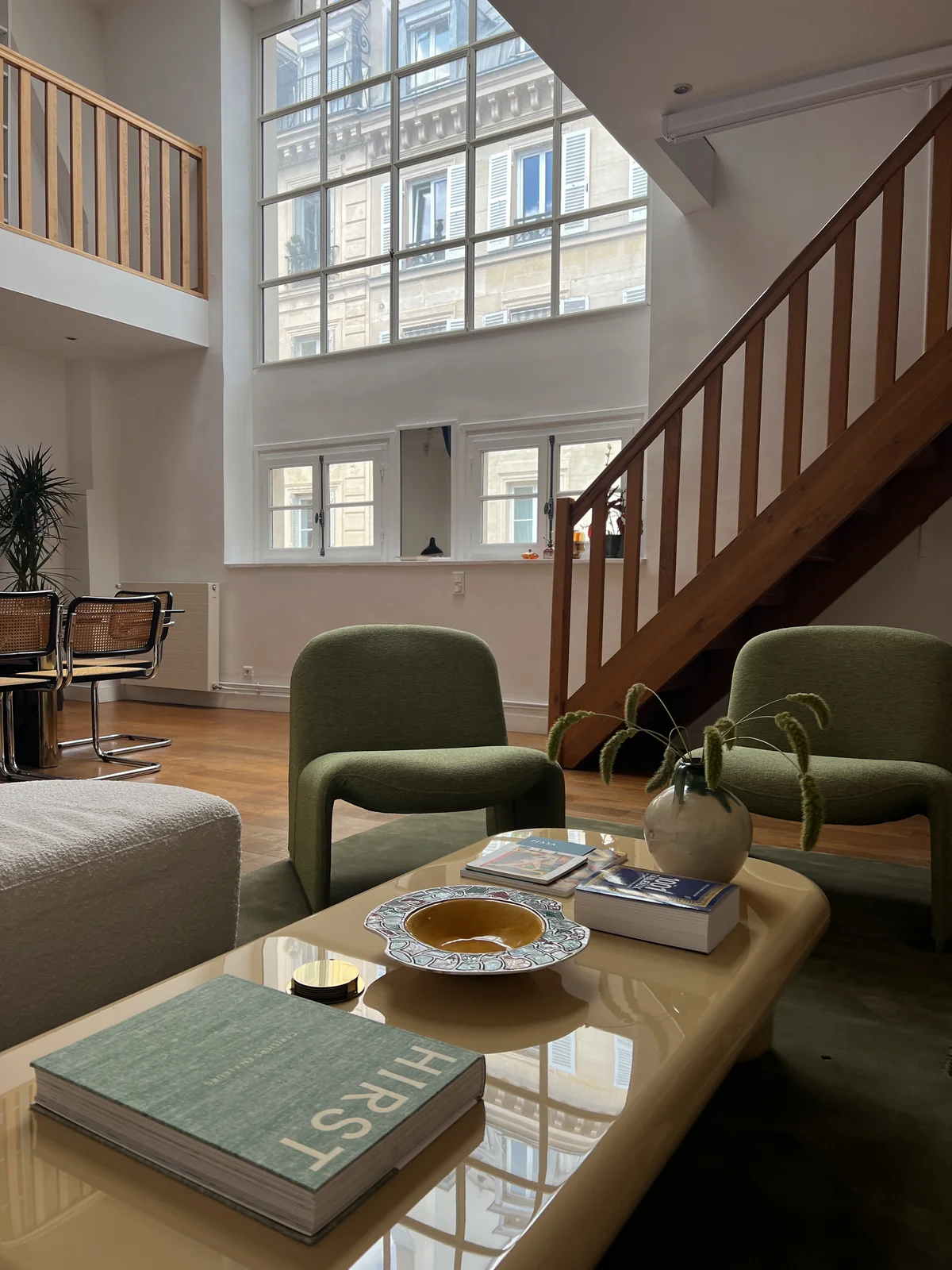 Green upholstered armchairs drawn around a low cream stone table holding art books, a glazed bowl and a spherical glass lamp, in a tall apartment whose paned windows look across to a carved stone facade; an open-tread wooden stair climbs the wall beside them.